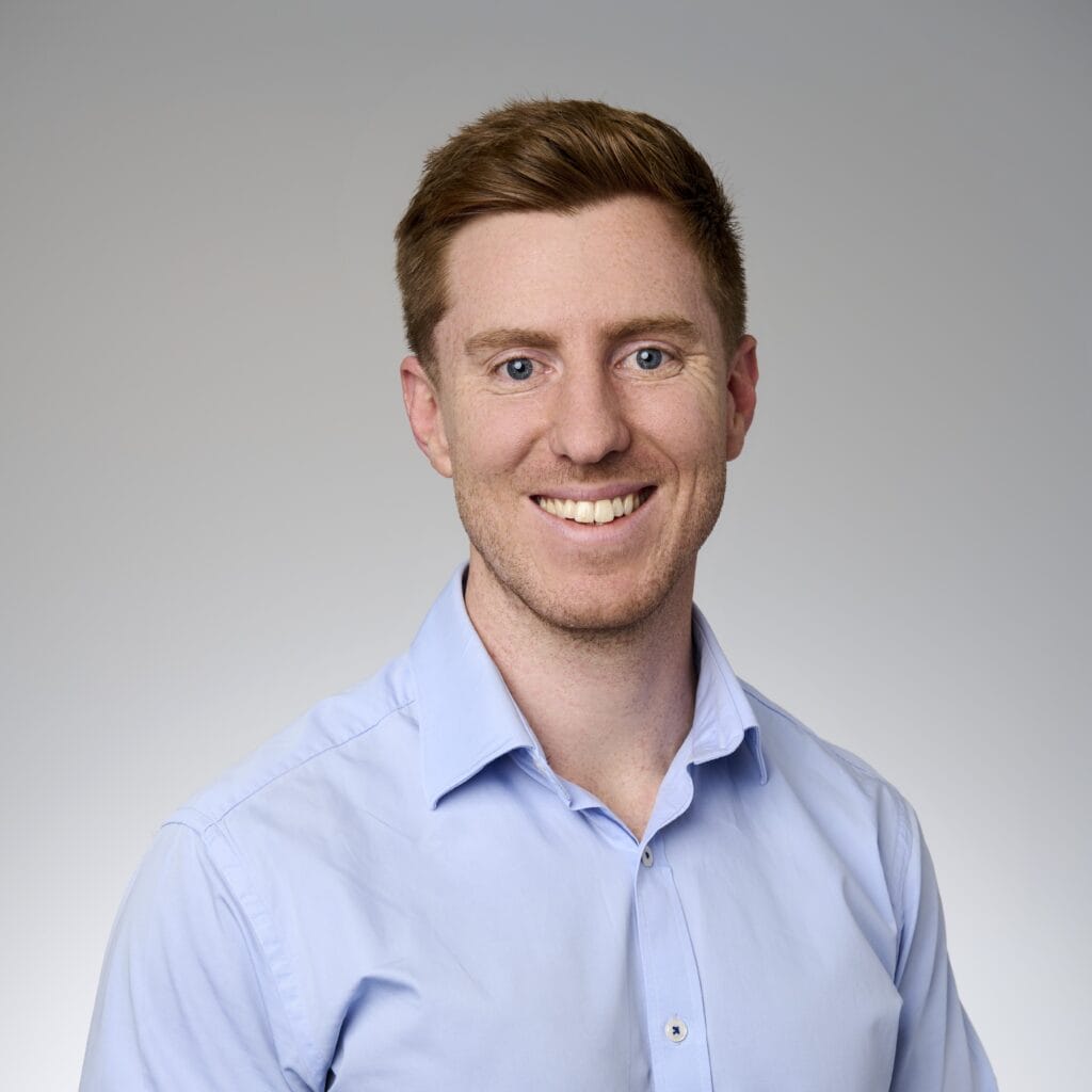 A corporate headshot of Sebastian Ryan, smiling and wearing light blue clothing.
