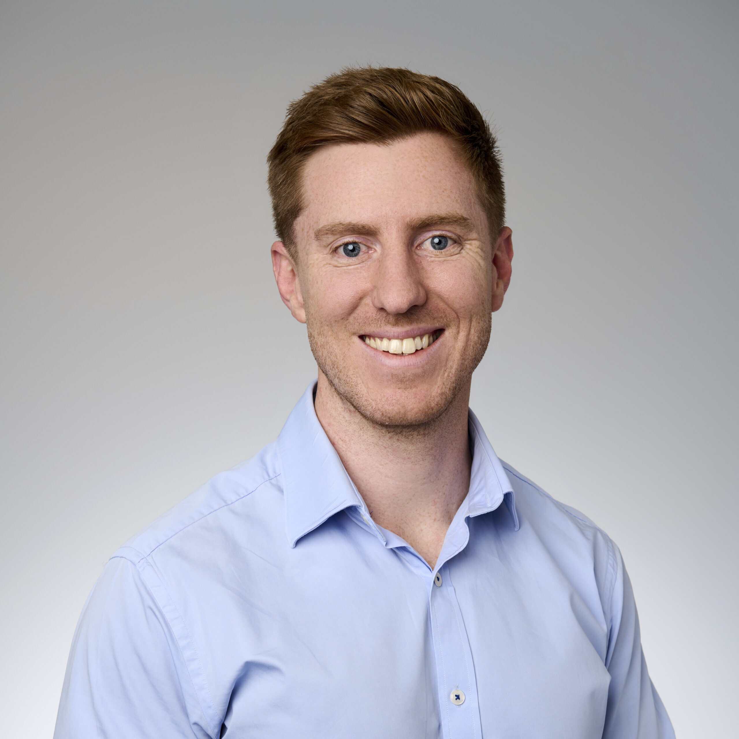 A corporate headshot of Sebastian Ryan, smiling and wearing light blue clothing.
