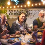 Happy family eating and drinking wine at barbecue dinner outdoor – Multiracial mature and young people having fun at bbq sunday meal – Food and summer lifestyle concept – Focus on asian woman face